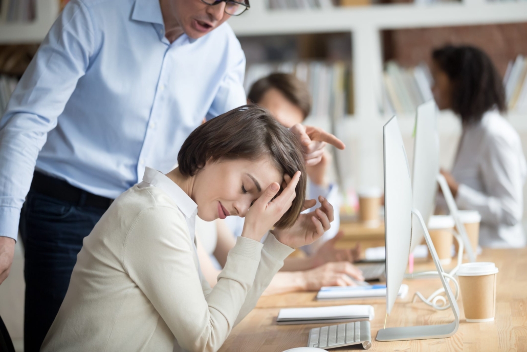 Woman looking down at her keyboard with eyes closed and hands up towards her forehead while a man behind her points at her screen and looks like he's reprimanding her